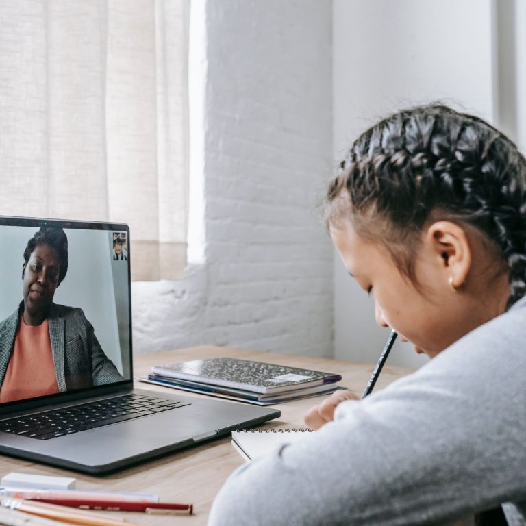 Side view of focused Asian schoolgirl sitting at wooden table and writing notes during remote lesson on laptop with African American teacher