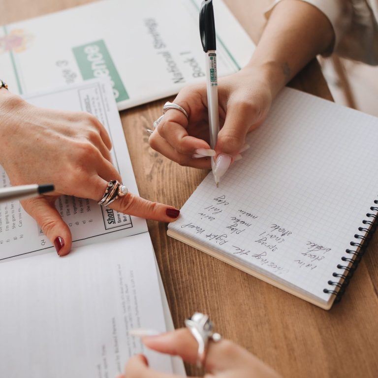 Close-up of hands writing in a notebook while studying documents on a wooden table.